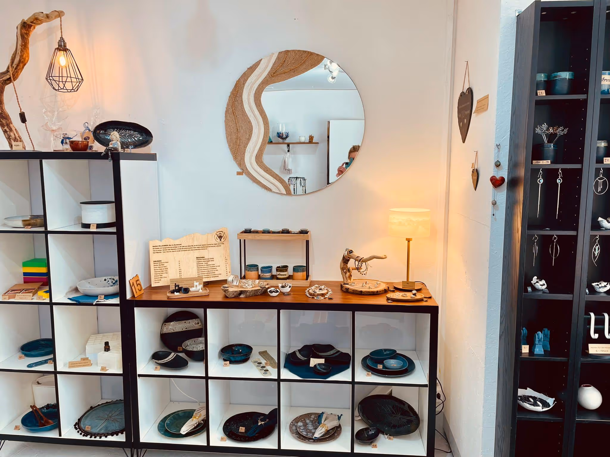Display of handcrafted ceramic plates, bowls, and jewelry arranged on white and black shelves under a round mirror with a wooden frame, lit by a table lamp and hanging light.