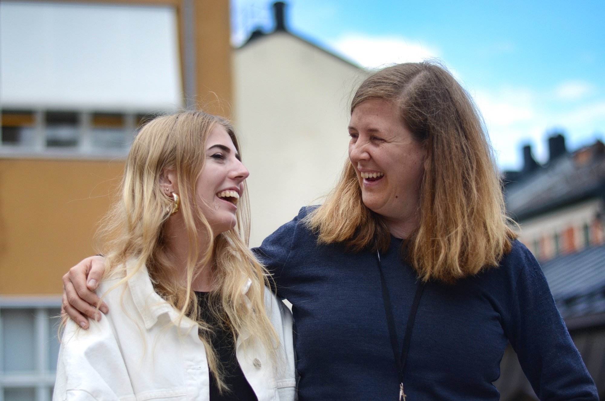 Två kvinnor står utomhus och skrattar tillsammans med armarna runt varandra.