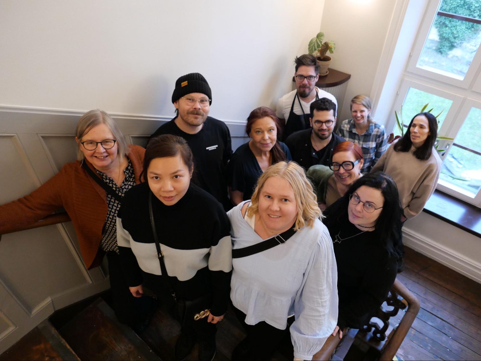 En grupp på elva personer som ler och står tätt tillsammans på en trappa inomhus nära ett fönster.