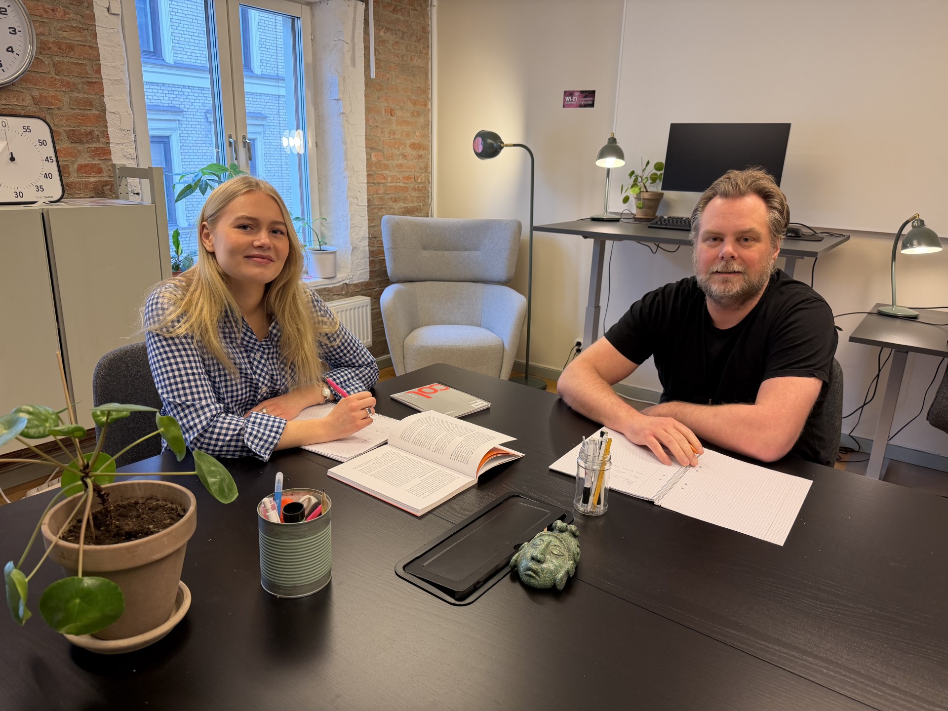 Två personer, en kvinna och en man, sitter vid ett bord med anteckningsböcker och pennor i ett kontorsrum med fönster, växter och datorskärmar i bakgrunden.