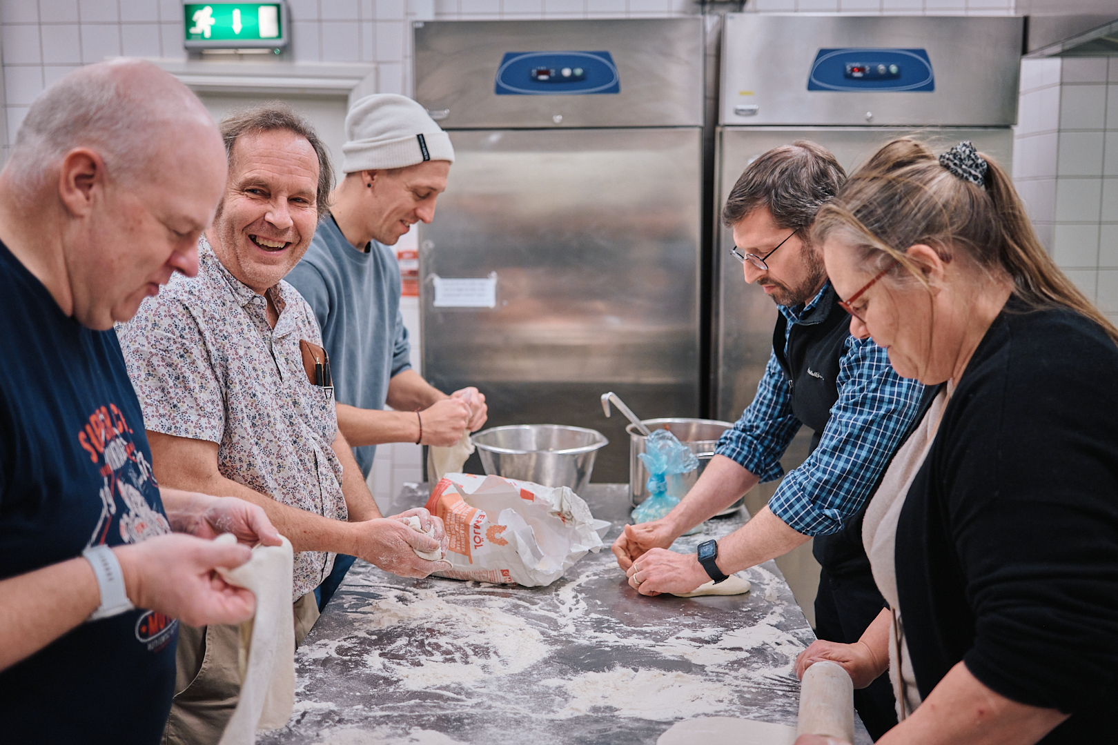 Fem personer bak ett bord i ett kök knådar och rullar ut deg med mjöl på bordet.