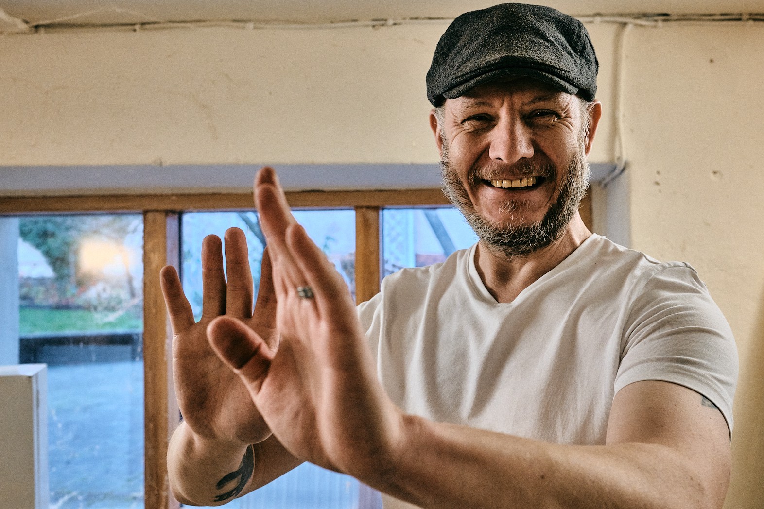 Leende man med keps och vit t-shirt som håller upp händerna framför sig inomhus.