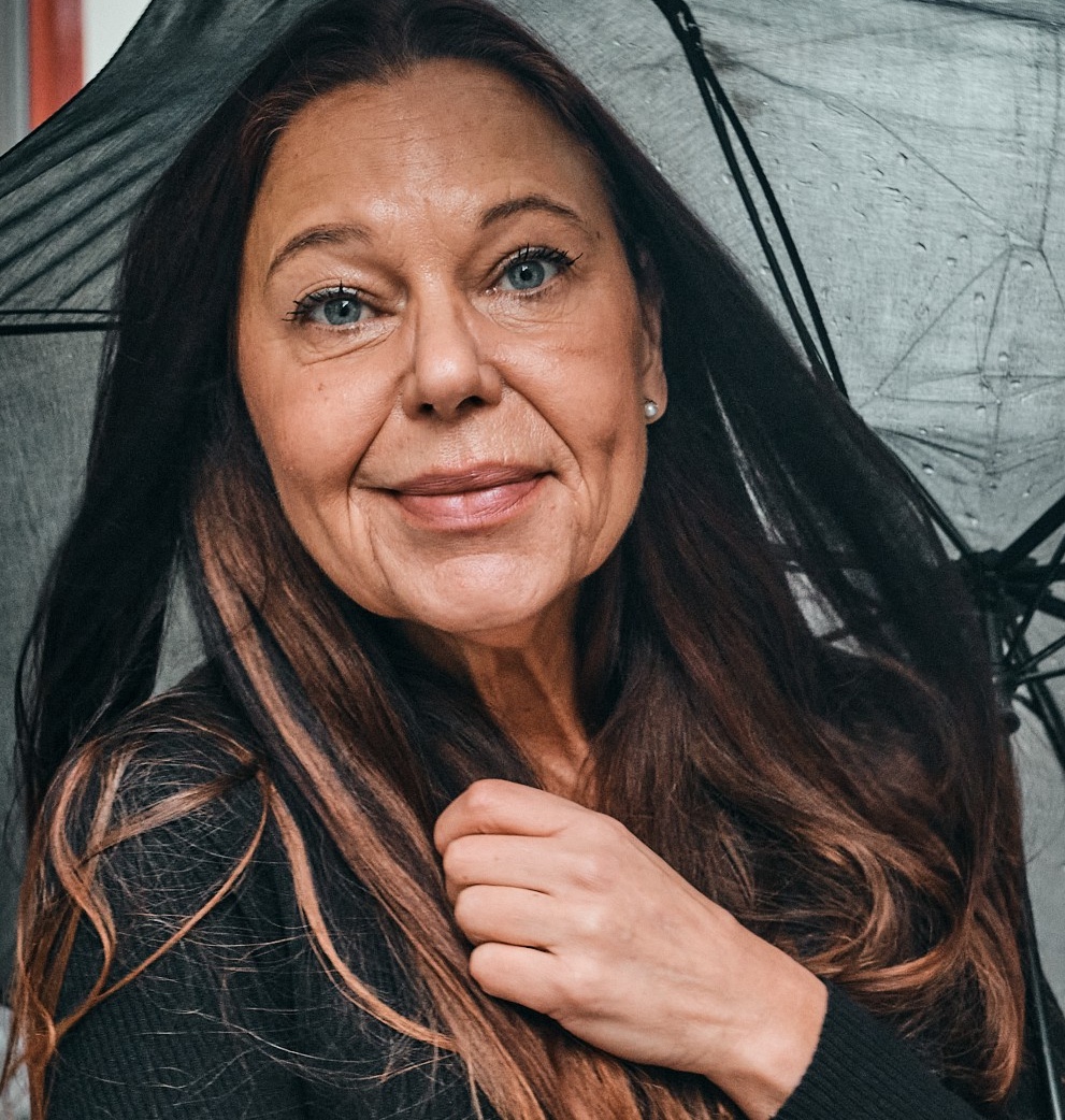 Närbild på leende kvinna med långt brunt hår som håller i ett svart paraply i regnet.