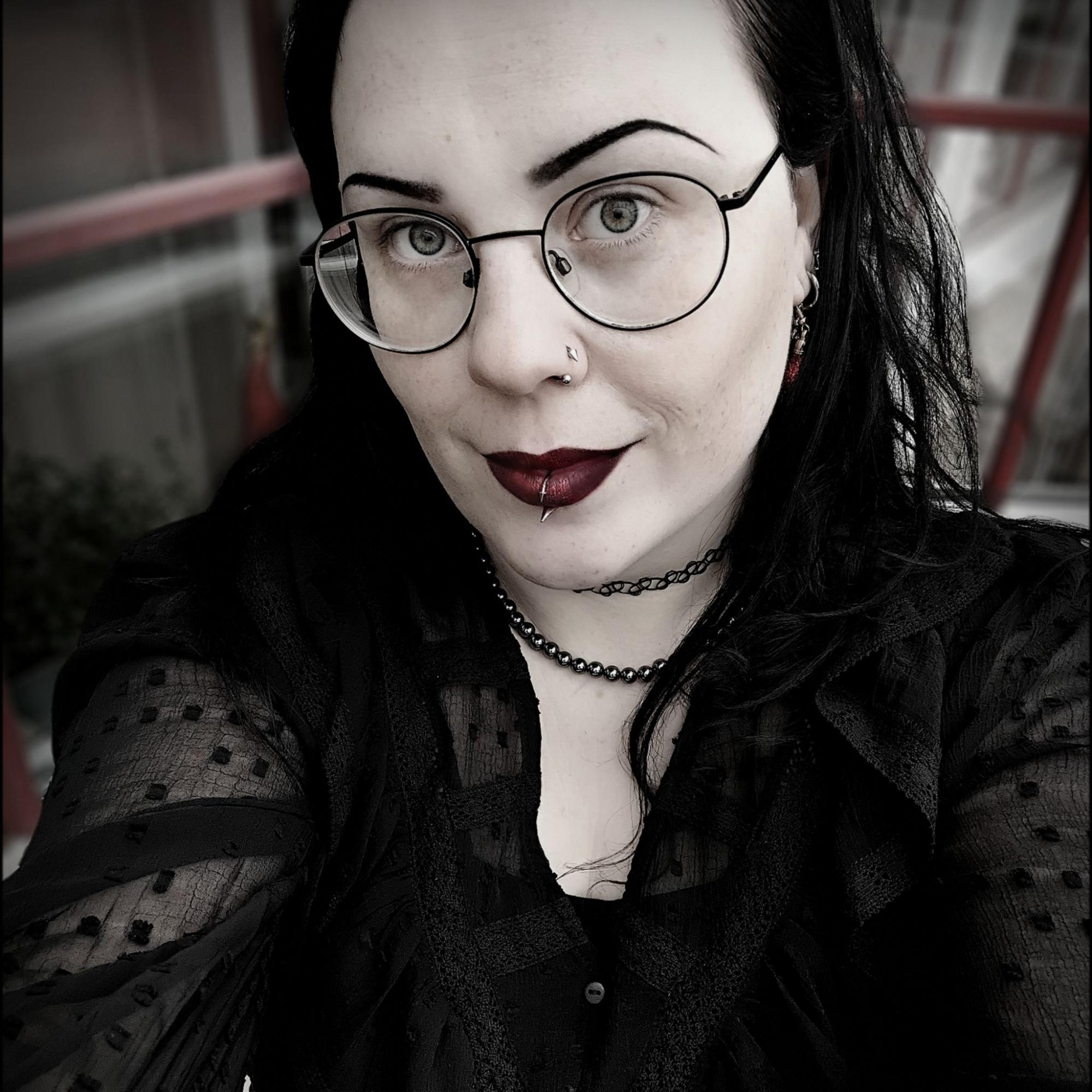 Närbild av kvinna med glasögon, mörkt hår, mörka kläder och flera piercingar i ansiktet, inklusive en läppiercing och näsring.