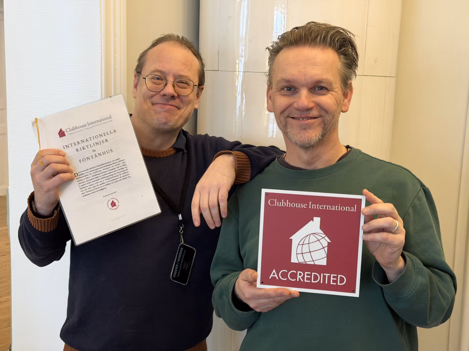 Två män ler och visar dokument, en håller en pärm med texten 'Internationella riktlinjer för fontänhus' och den andra håller en skylt med 'Clubhouse International Accredited'.