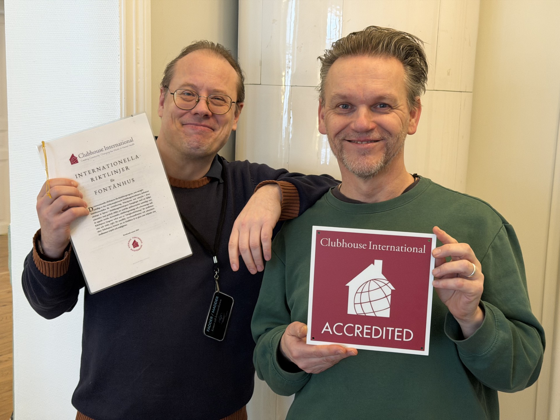 Två män ler och visar dokument, en håller en pärm med texten 'Internationella riktlinjer för fontänhus' och den andra håller en skylt med 'Clubhouse International Accredited'.