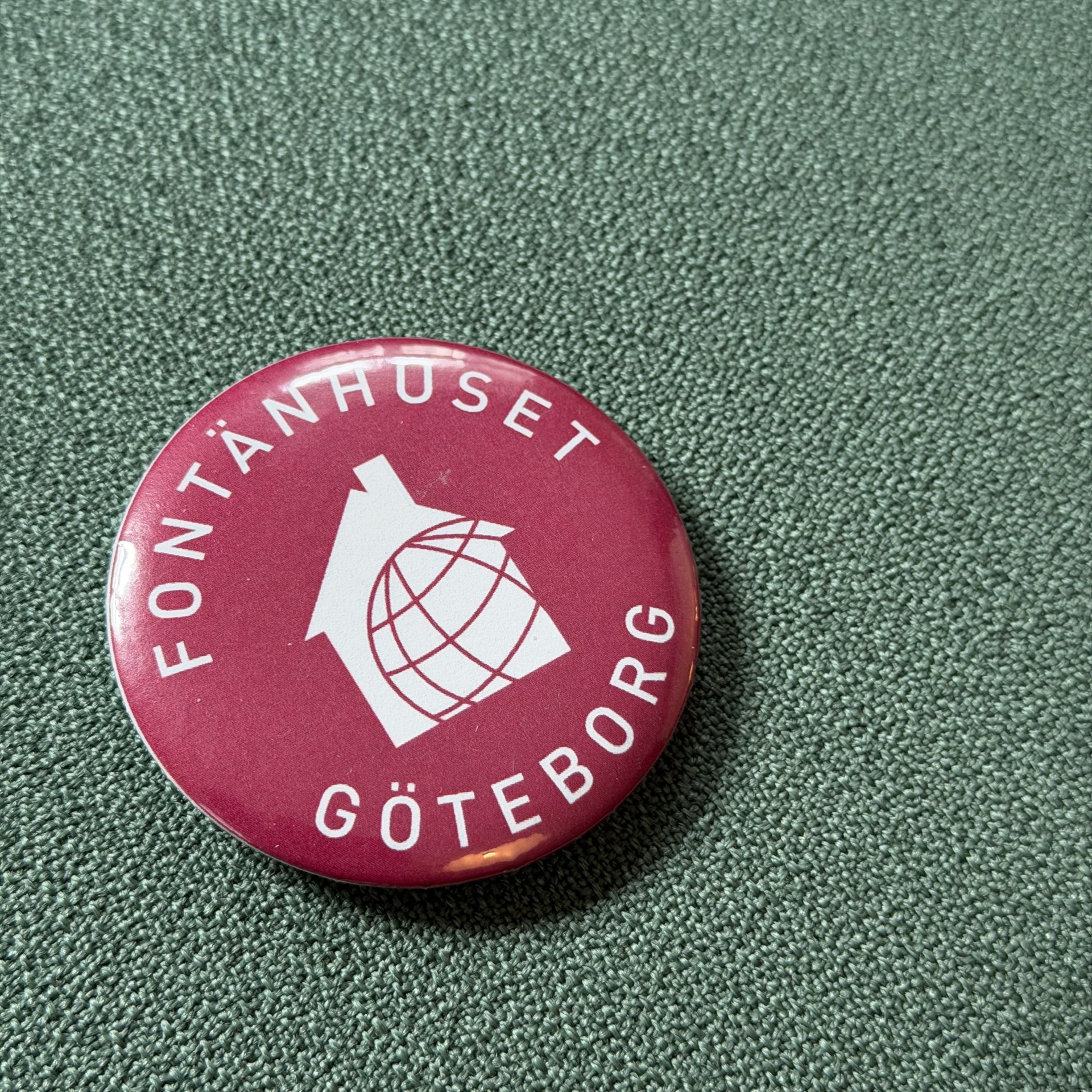 Rund knapp med texten Fontänhuset Göteborg och en vit ikon av ett hus med en stiliserad glob på röd bakgrund.