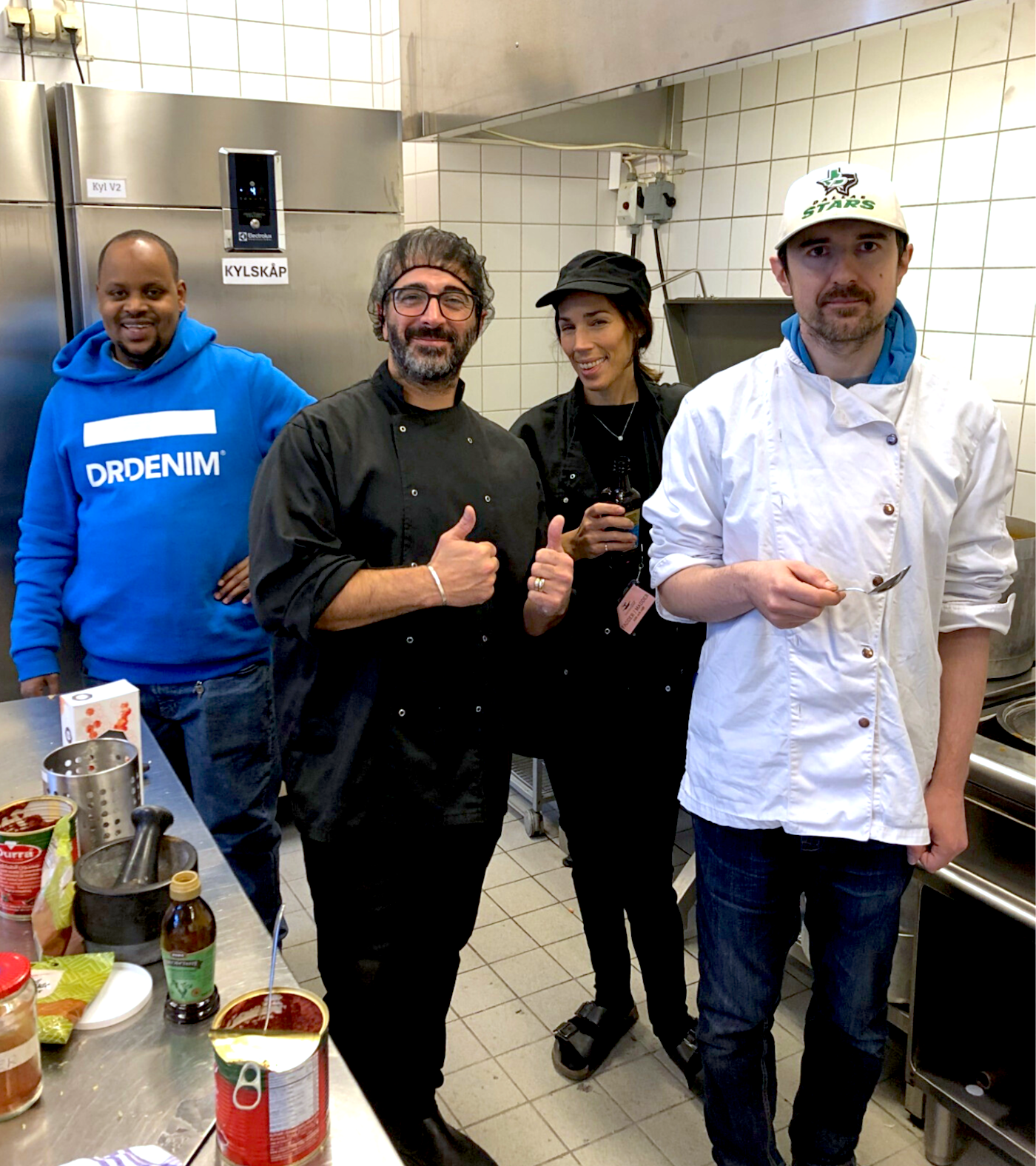 Fyra personer i ett kök, två av dem i svarta kockjackor, en man i blå huvtröja och en i vit kockjacka med en sked i handen.