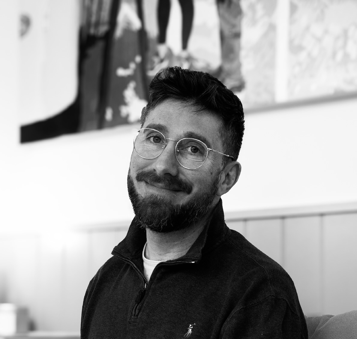 Smiling man with glasses and beard wearing a dark quarter-zip sweater seated indoors.