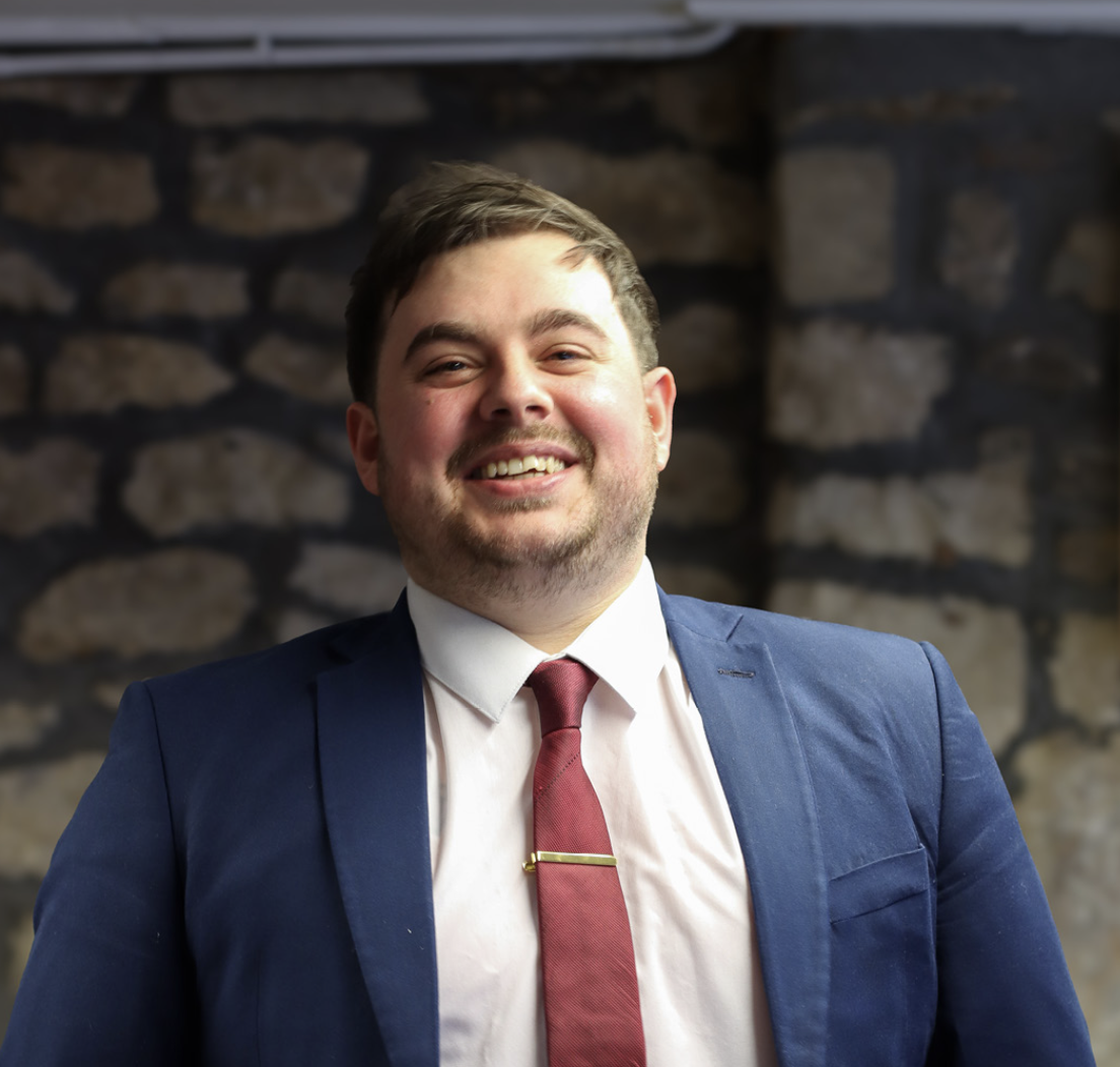 Smiling man with dark hair wearing a blue suit jacket, white shirt, and red tie standing in front of a stone wall.