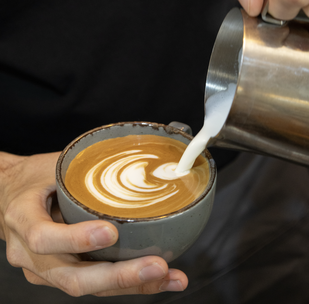 Hand holding a ceramic cup while milk is being poured to create latte art on coffee.