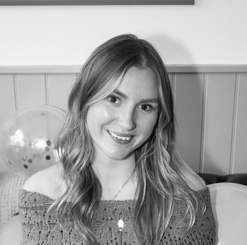 Smiling young woman with long blonde hair, wearing a dark sweater and a pendant necklace, seated indoors.