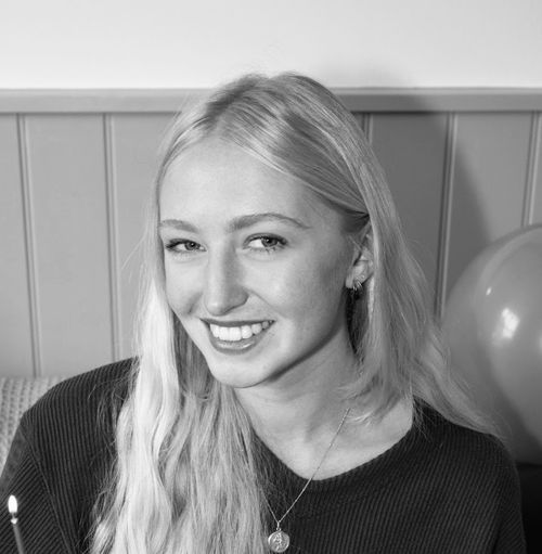 Smiling young woman with long blonde hair wearing a dark sweater and a pendant necklace.