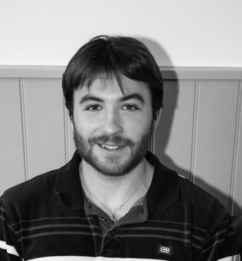 Smiling man with short dark hair and a beard, wearing a striped polo shirt, sitting against a paneled wall.