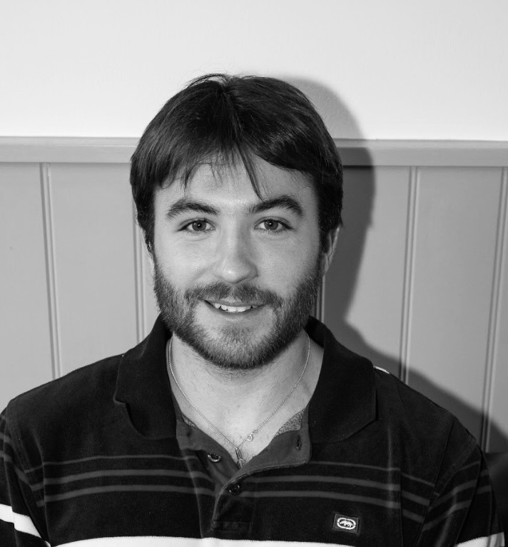 Smiling man with short dark hair and a beard, wearing a striped polo shirt, sitting against a paneled wall.