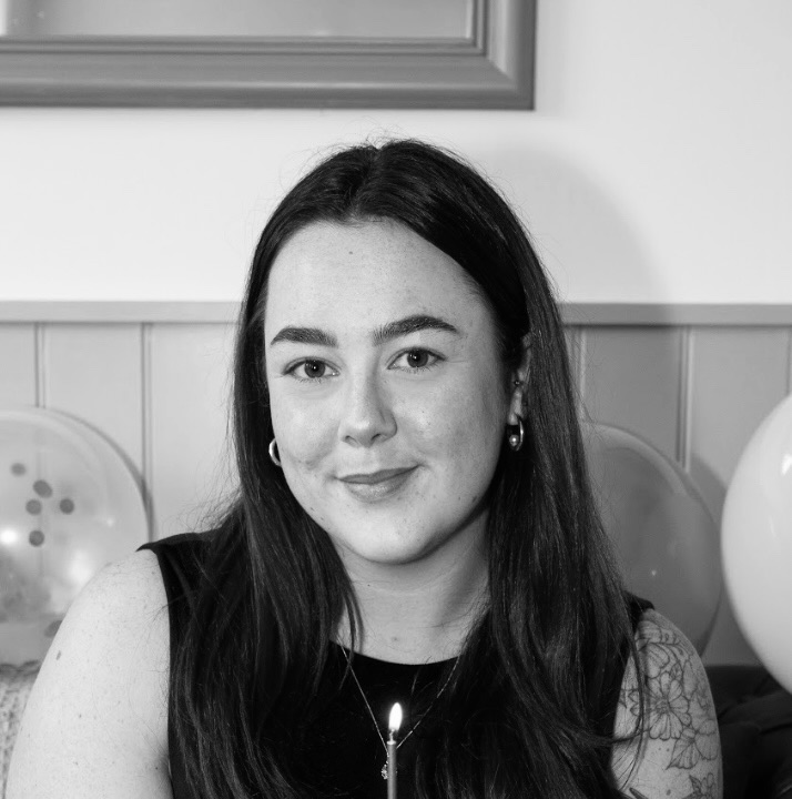Black-and-white portrait of a smiling young woman with long dark hair, hoop earrings, and floral tattoos on her left arm, sitting indoors with balloons in the background.