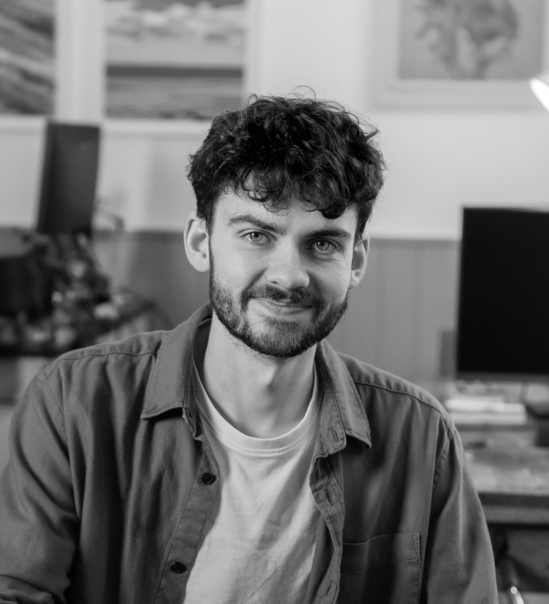 Smiling young man with curly hair and beard wearing a button-up shirt over a t-shirt in a casual indoor setting.