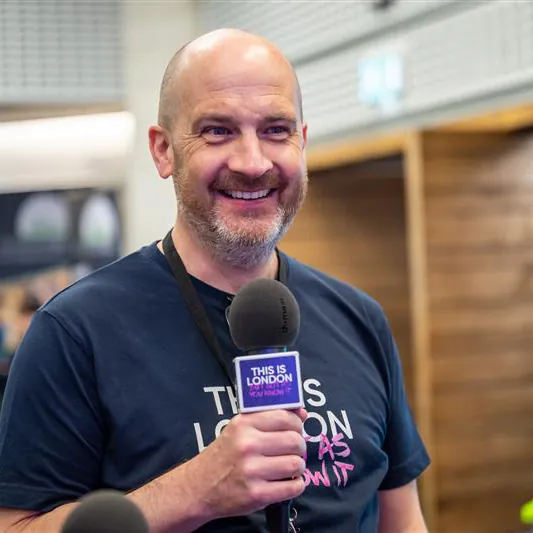 Kevin Baulf, Head of Sales. A smiling man holds a microphone with a This is London label. He wears a navy blue shirt and has a lanyard around his neck. The background is blurred, showing an indoor setting.