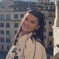Abbie Carlton, Digital Marketing Officer. A woman with long dark hair smiles at the camera while standing on a balcony, with sunlit buildings and a cityscape in the background.