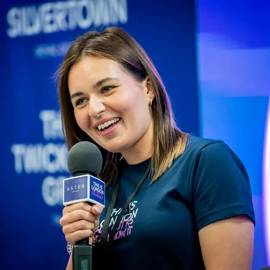 Cheryl Gibbens, Digital Marketing Manager. A woman with straight brown hair smiles while holding a microphone. She wears a navy blue T-shirt and stands in front of a blurred blue background with white text.