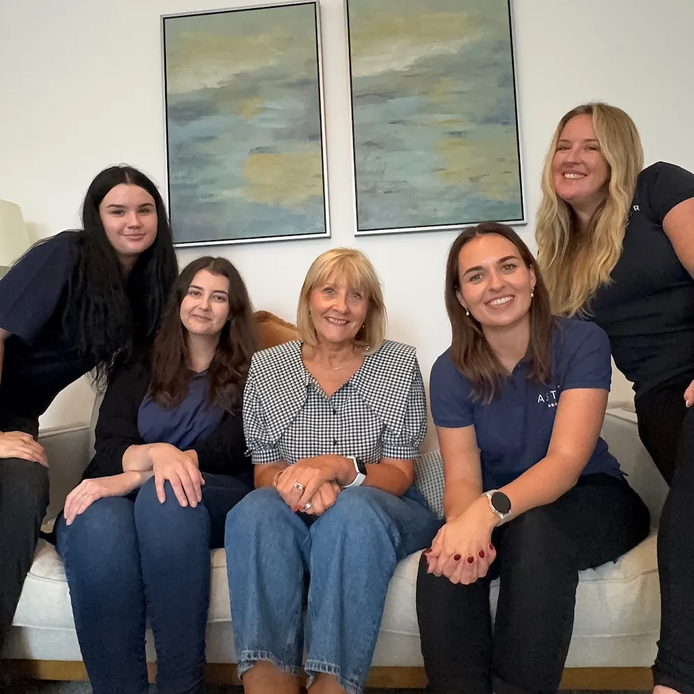 Nicci Stone, Sales Negotiator. Five women sit together on a sofa, smiling at the camera. Two abstract paintings hang on the wall behind them. The setting appears to be a cosy, well-lit living room.
