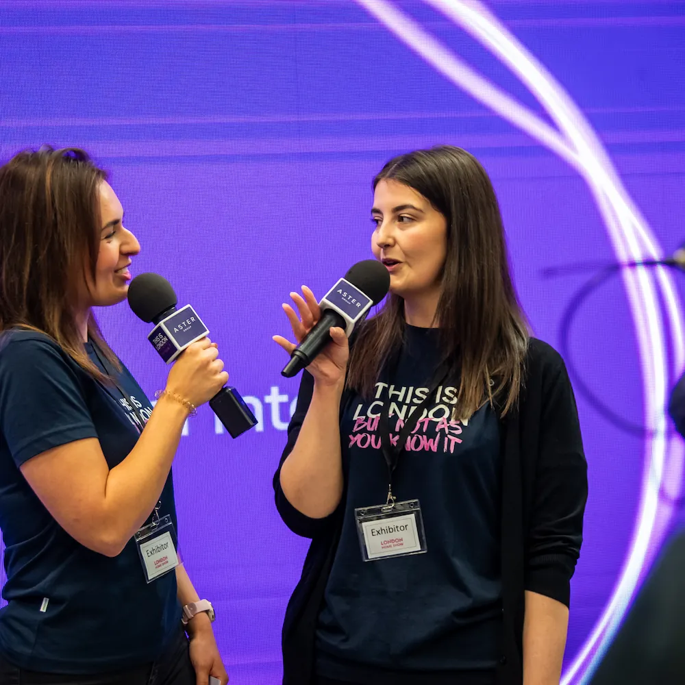 Chantelle Botwright, Marketing Officer. Two women wearing name badges and holding microphones speak on stage against a vibrant blue and purple background with white light streaks. Both are smiling and appear to be participating in an event or interview.