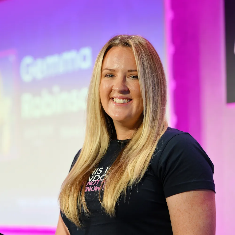 Gemma Robinson, Head of Marketing. A woman with long blonde hair smiles while standing in front of a purple and pink background with blurred text. She wears a navy T-shirt.