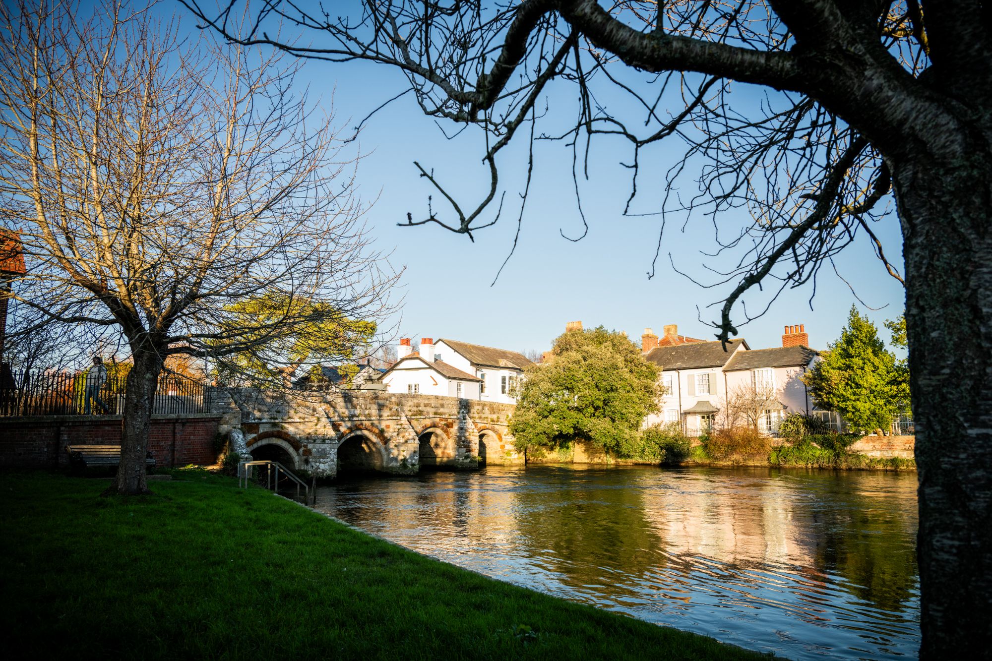 A stone bridge spans a calm river, with white houses and green shrubs nearby. Leafless trees frame the scene under a clear blue sky.