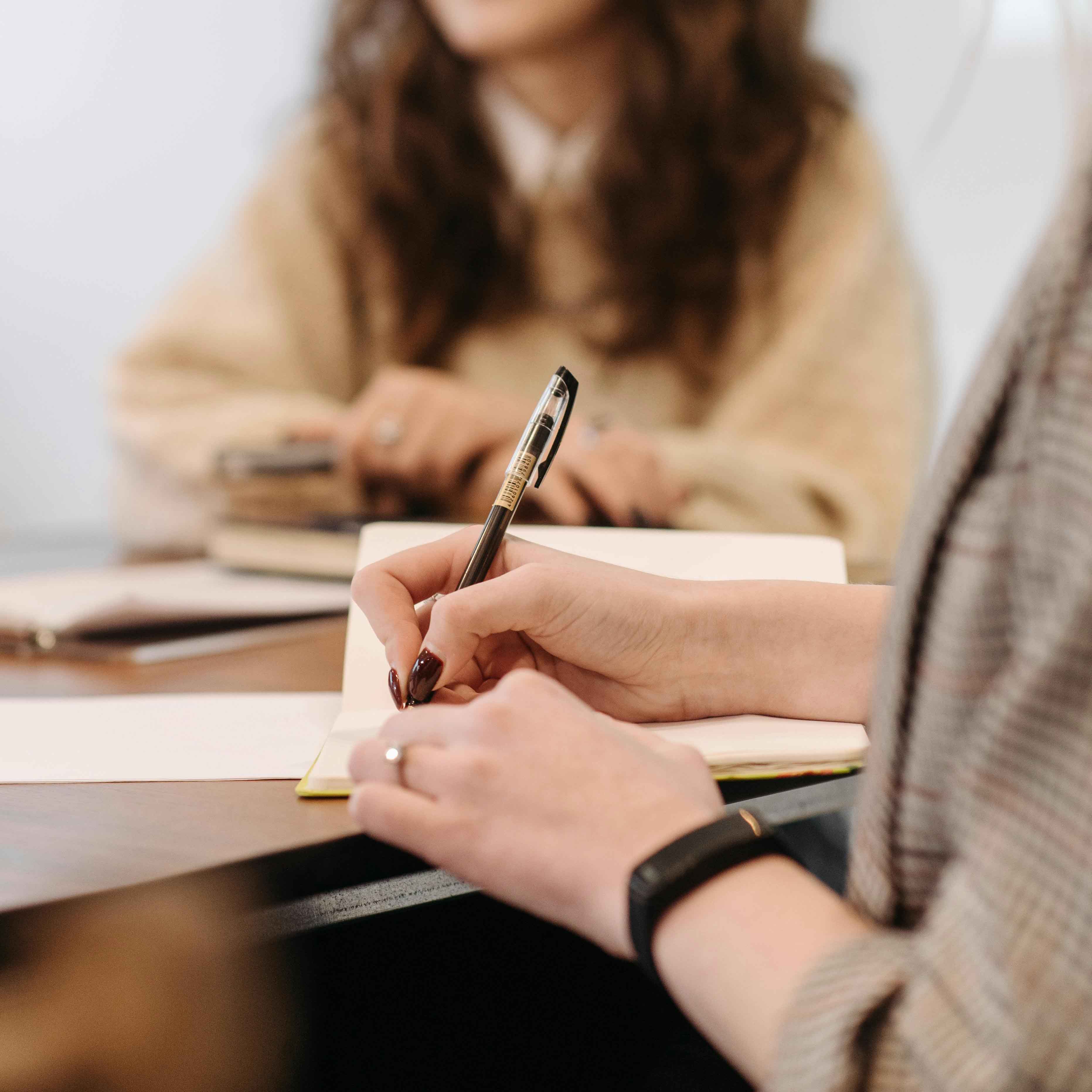 Hand mit schwarzem Stift schreibt in ein offenes Notizbuch während eine weitere Person im Hintergrund sitzt.
