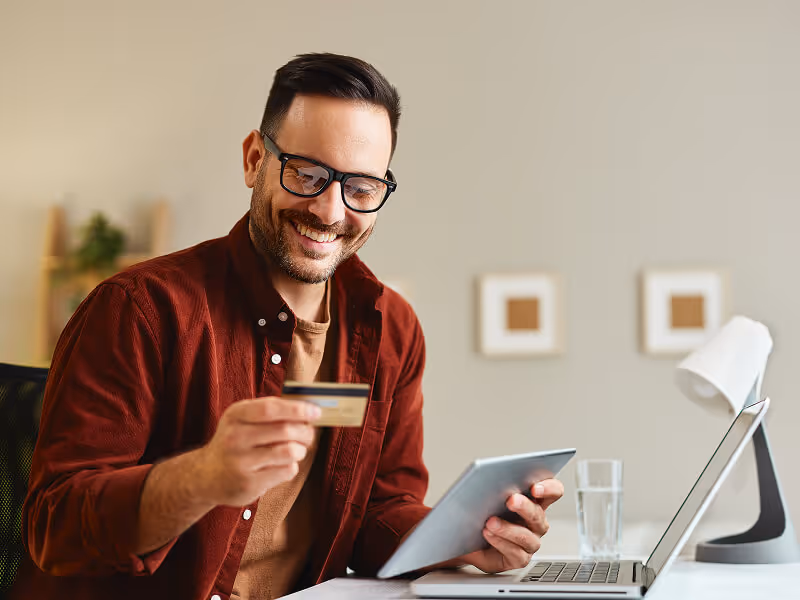 Smiling man wearing glasses holding a credit card and a tablet while sitting at a desk with a laptop and a glass of water.