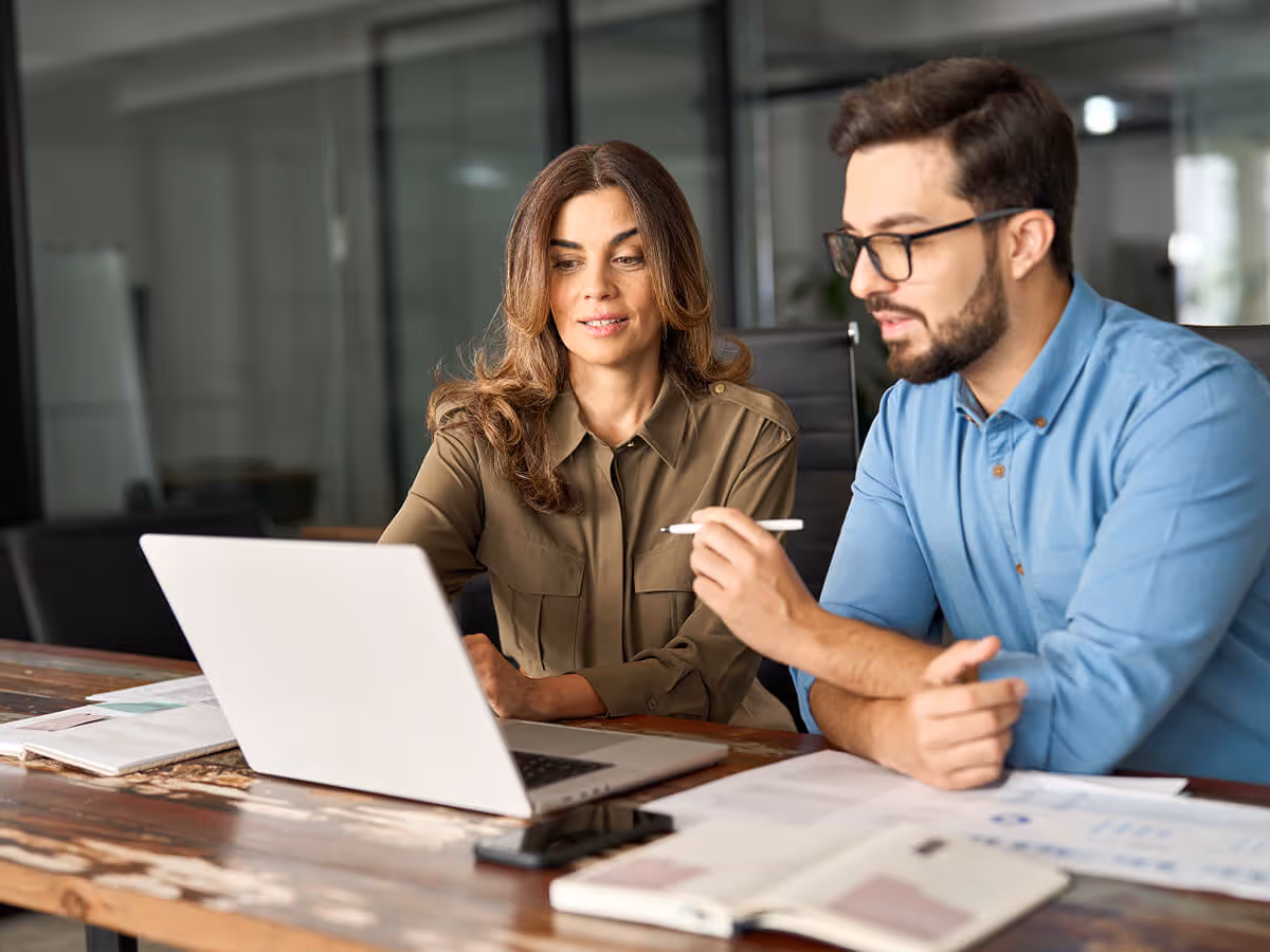 A woman and a man working together on a laptop at a wooden table in an office.