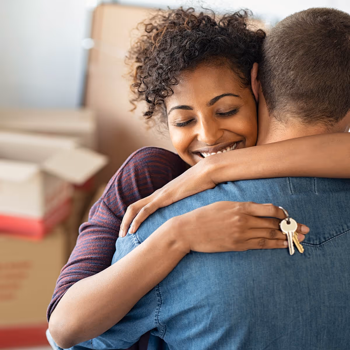 Smiling woman hugging a man while holding a set of home keys, with moving boxes in the background.