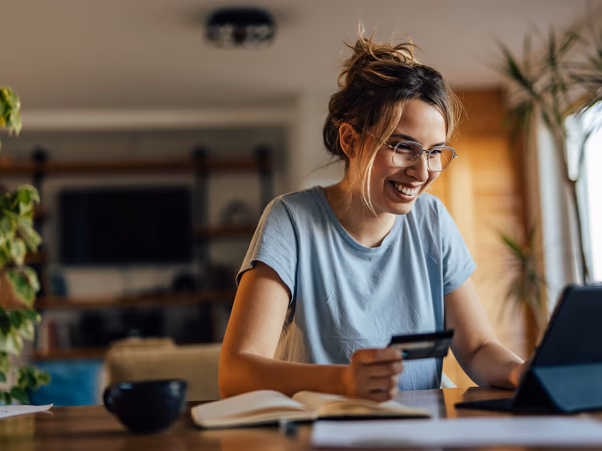 Smiling woman with glasses holding a credit card and looking at a tablet in a cozy home setting.