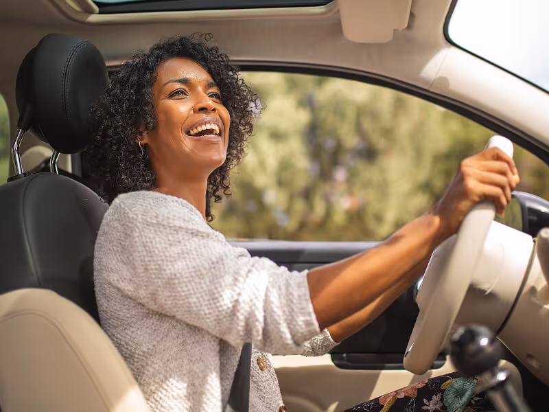 Smiling woman with curly hair driving a car, view from the side seat showing her holding the steering wheel.