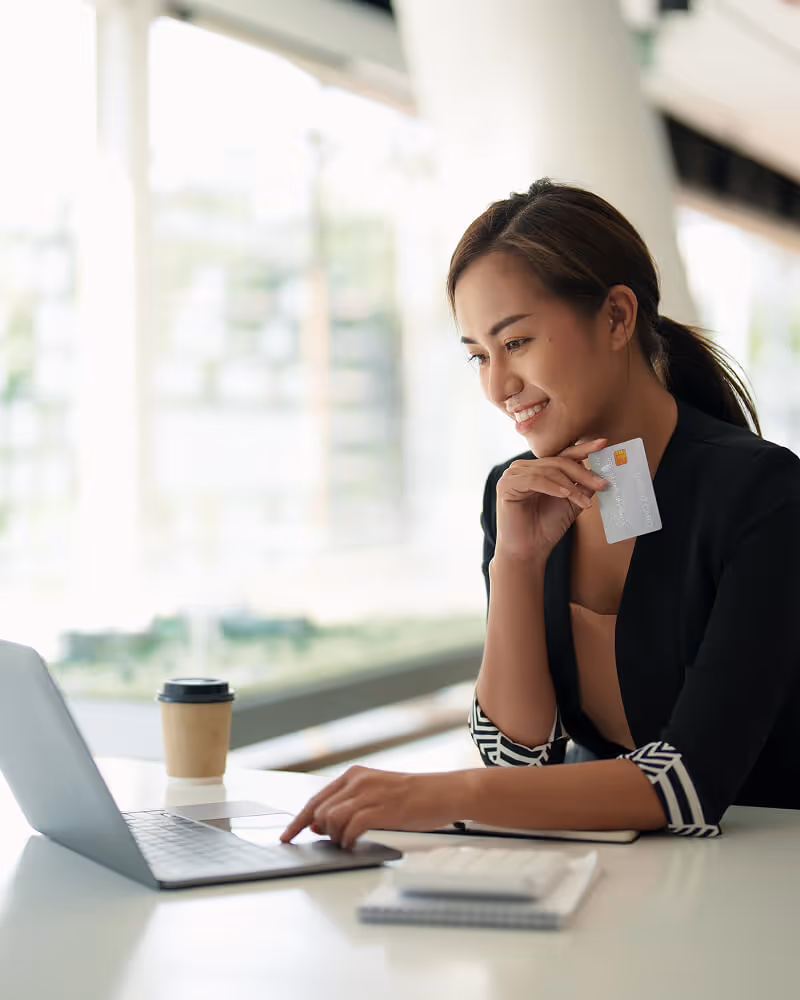 Smiling woman holding a credit card and using a laptop with a coffee cup nearby.