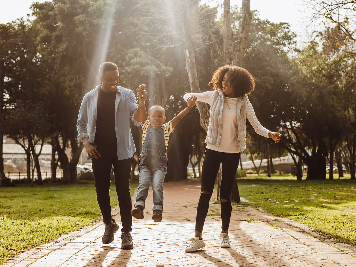 Smiling parents swinging their young child by the arms while walking on a sunlit park path.