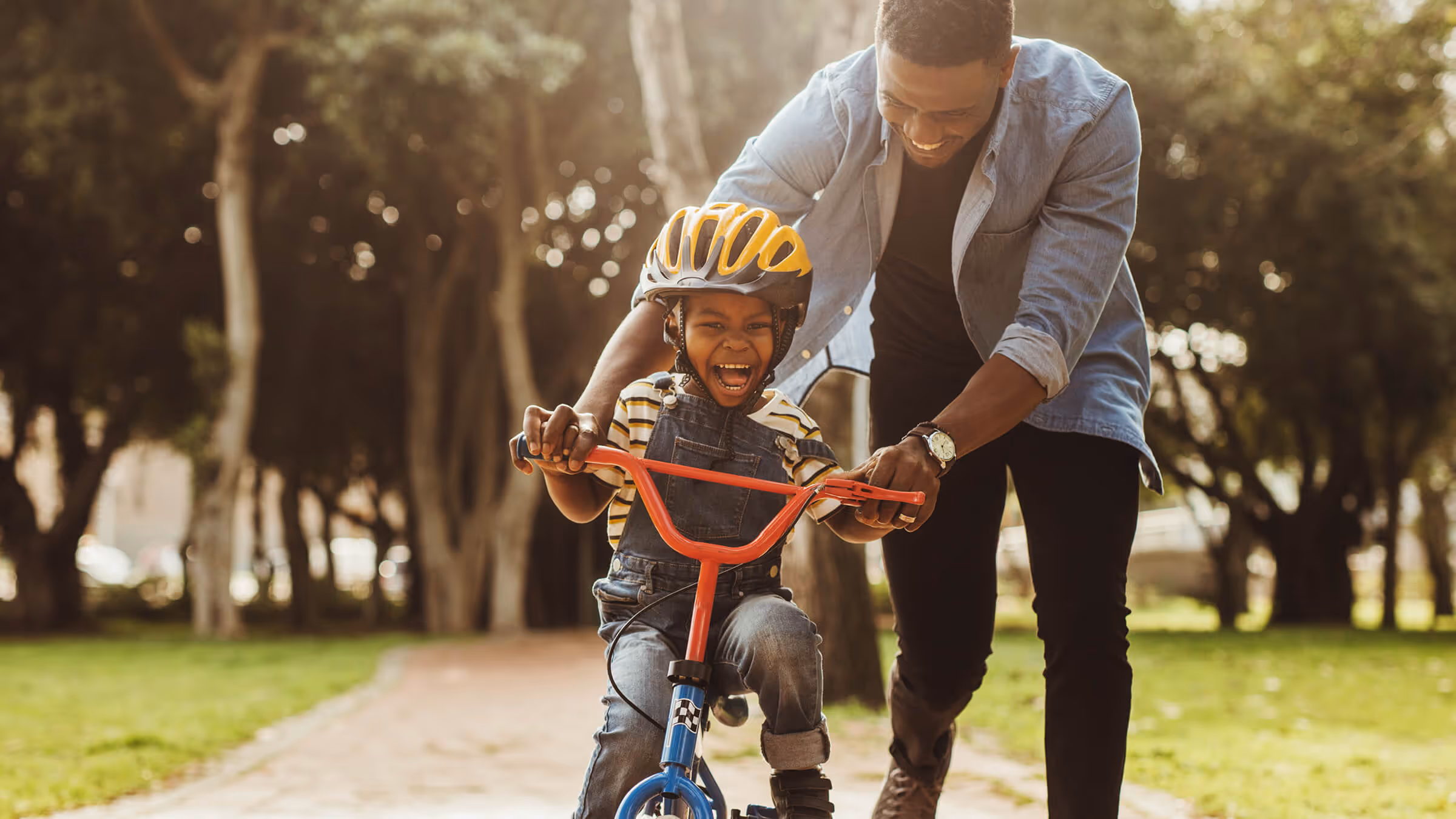 Smiling father helping his excited child ride a bicycle on a park path.