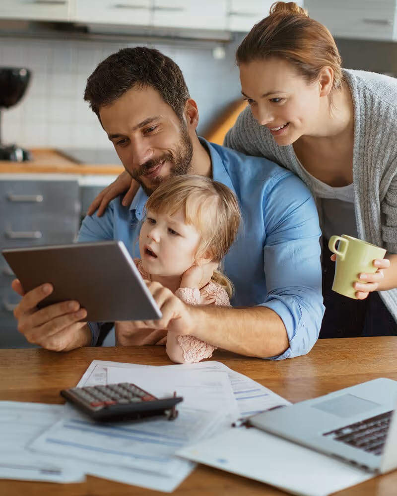 Parents and their young daughter looking at a tablet together at a table with documents, a calculator, and a laptop.