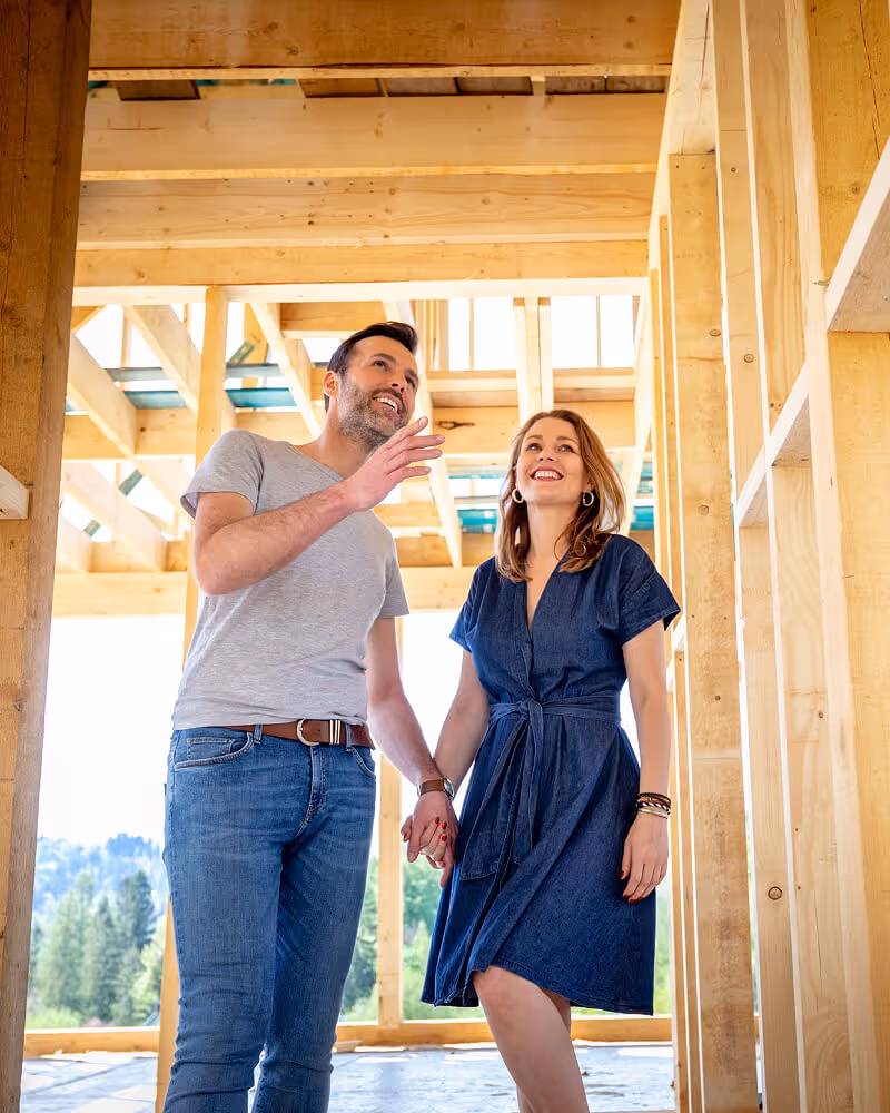 Smiling couple holding hands inside a wooden house frame under construction.