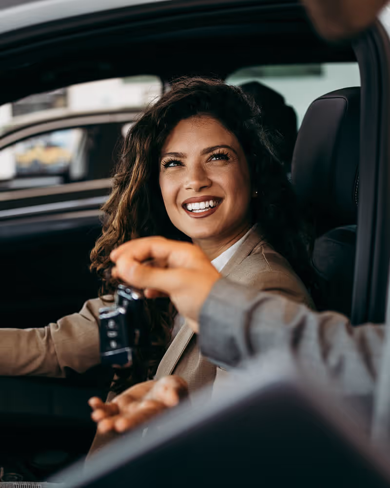 Smiling woman sitting inside a car receiving keys from a person outside.