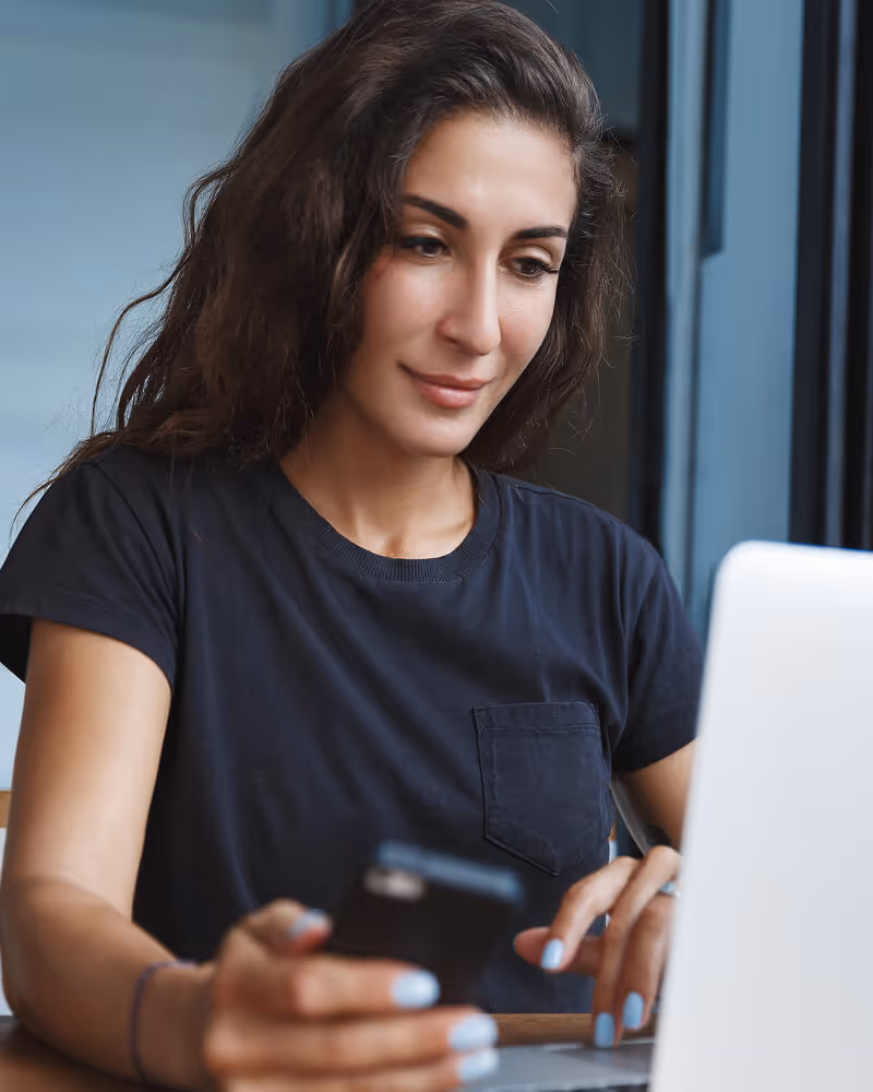 Woman with long dark hair using a smartphone and laptop at a wooden table.