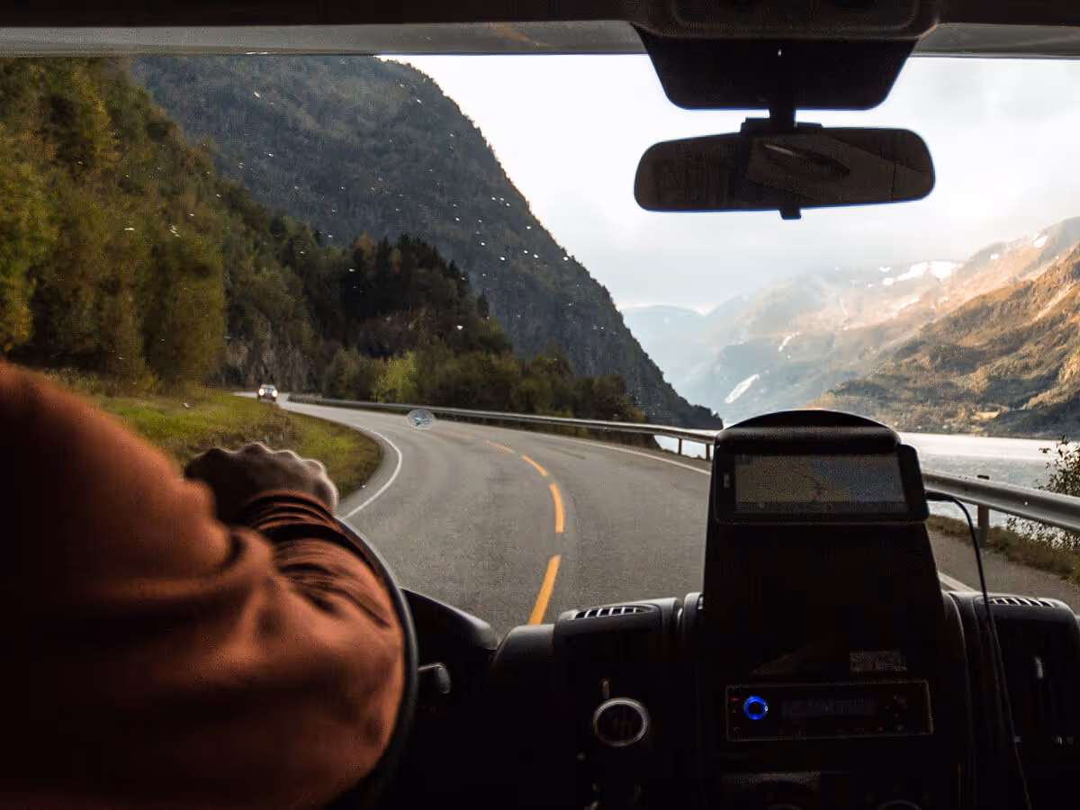 View from inside a vehicle driving on a winding road beside a lake with mountains in the background.