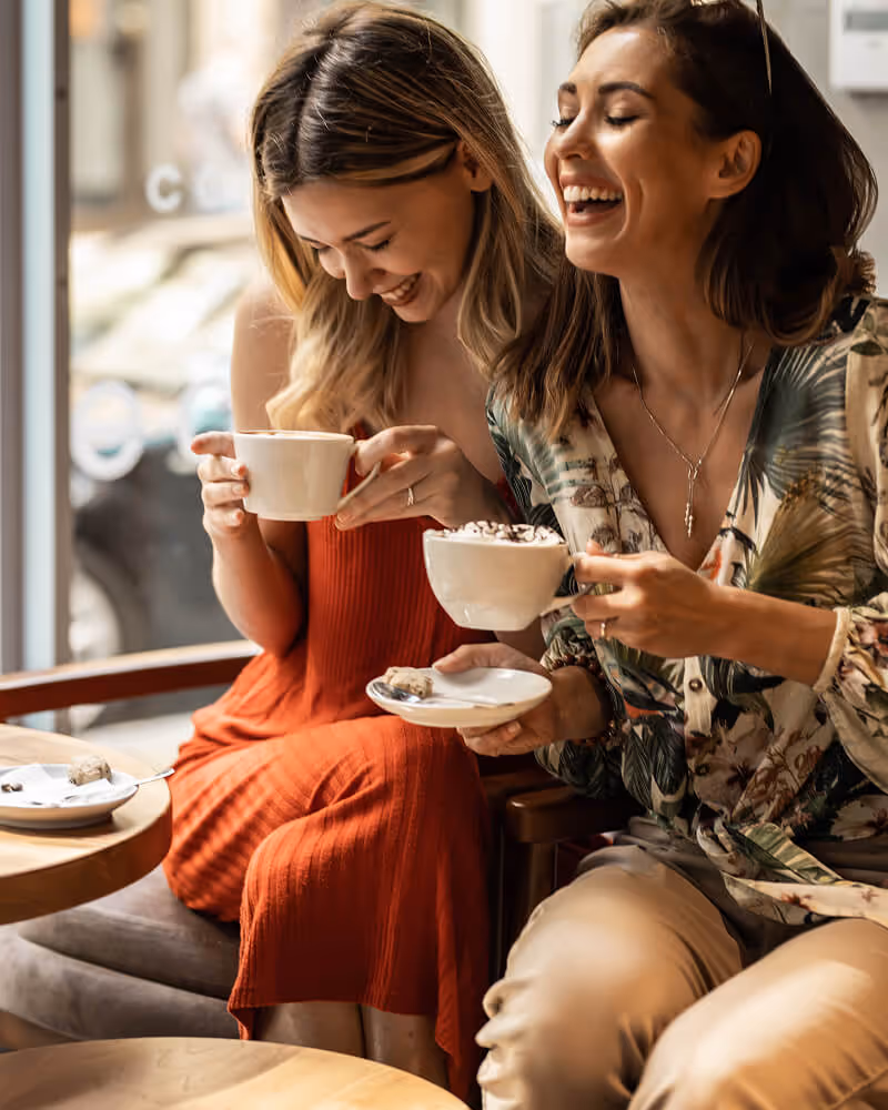 Two women laughing together while sitting in a cafe, each holding a cup of coffee with a saucer.