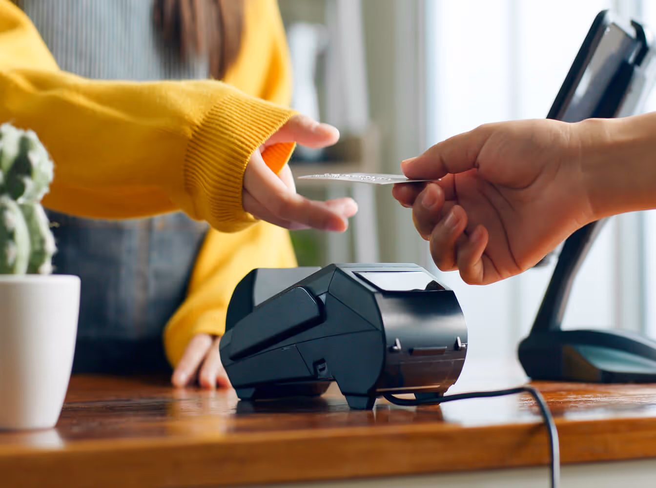 Customer in yellow sweater handing a debit card to a cashier with a card payment terminal on the counter.