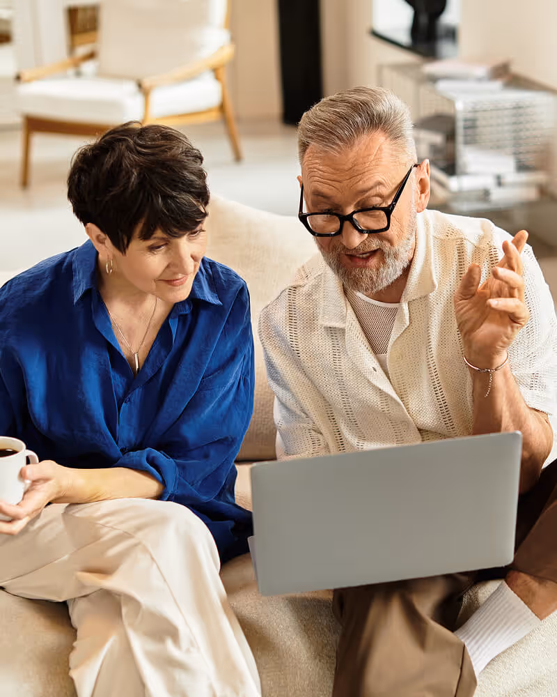 Older man and woman sitting on a couch looking at a laptop, with the woman holding a cup.