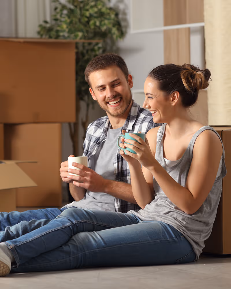 Smiling young couple sitting on the floor with mugs, surrounded by moving boxes.