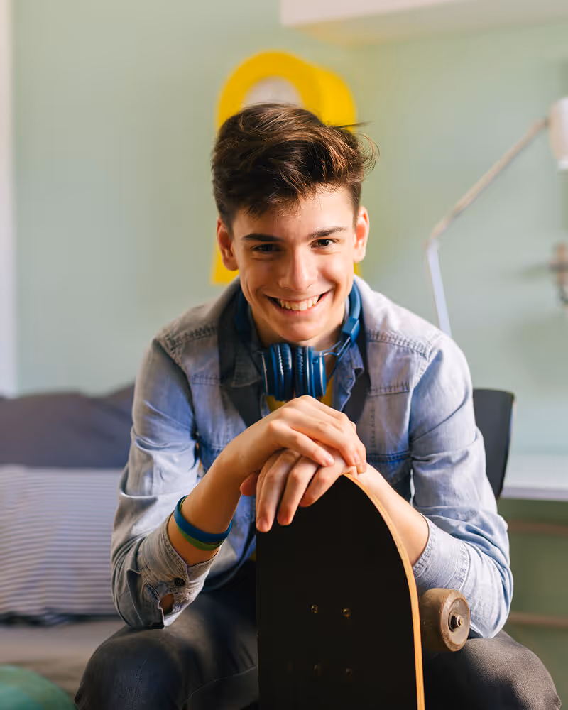 Smiling teenage boy sitting indoors holding a skateboard with headphones around his neck.
