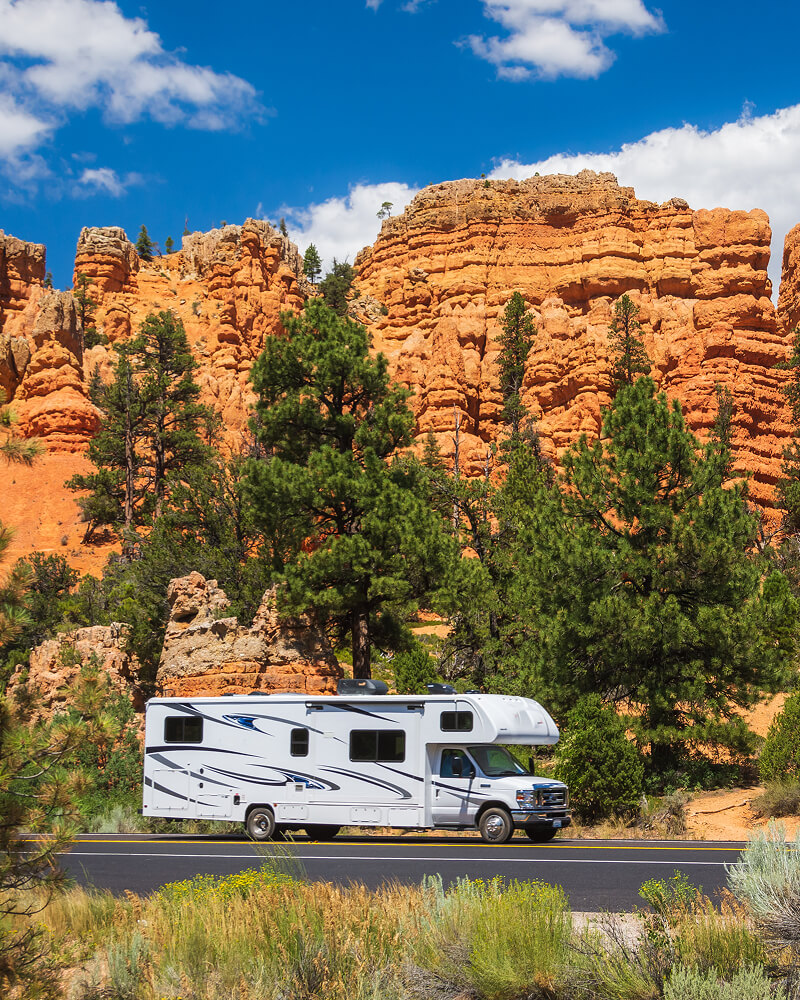 White RV driving on a paved road with green pine trees and red rock cliffs in the background under a partly cloudy blue sky.