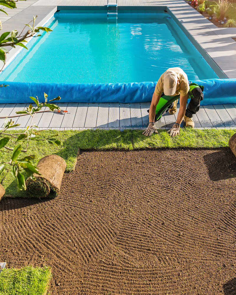 A gardener wearing gloves and a cap laying fresh sod near a swimming pool with blue water and a rolled-up pool cover.