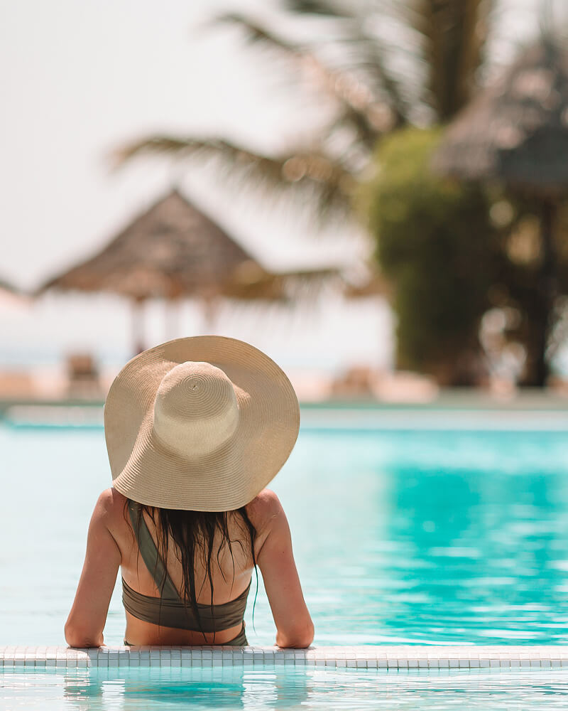 Woman wearing a wide-brimmed hat relaxing by a clear blue pool with palm trees and thatched umbrellas in the background.